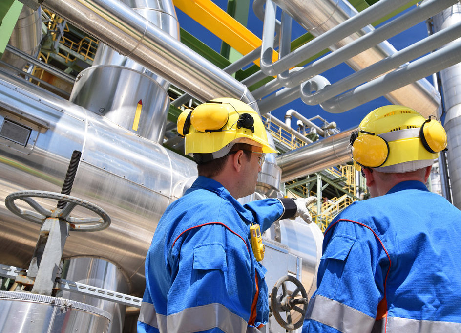 Zwei Arbeiter in blauen Overalls mit gelben Schutzhelmen und darauf festgeklemmten Kopfhörern schauen auf eine Vielzahl von Rohren in einem Werk und diskutieren darüber